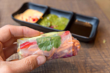 Hand holds a summer roll filled with avocado, mango, paprika, red cabbage, carrots and rice noodles with various sauces in the background - image gratuit #460977 